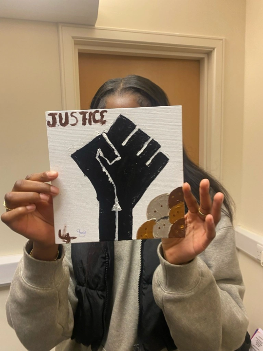 Person holding a square canvas in front of their face. The canvas depicts a closed fist pointing upwards and the word 'justice' on top left.