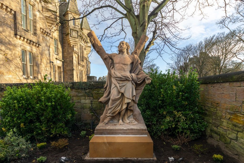 Statue of a man with cloth draped around his body and a cross behind him. The statue is on a pedestal and there are hedges, buildings and trees in the background.